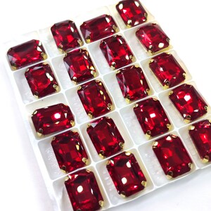 Puede incluir: Una bandeja de pedrería rectangular, facetada, de color rojo rubí, con engastes de metal dorado. Las gemas están dispuestas en un patrón de cuadrícula, cada una reflejando la luz. Ideal para manualidades, bisutería o adornos.