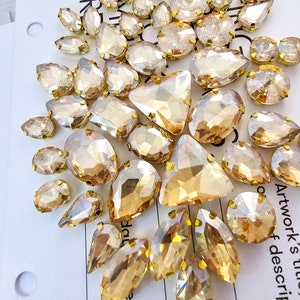 Puede incluir: Una colección de pedrería de color ámbar de varias formas, con engastes de metal dorado. Las piedras están dispuestas sobre una superficie blanca, mostrando diferentes cortes como lágrimas, óvalos y triángulos. La luz captura las facetas, creando un efecto brillante.