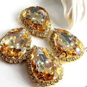 Pode incluir: Quatro grandes strass em forma de gota, com um tom amarelo dourado. Cada pedra é emoldurada por uma armação de metal dourado, adornada com pequenos strass transparentes. As gemas facetadas captam a luz, criando um efeito cintilante.
