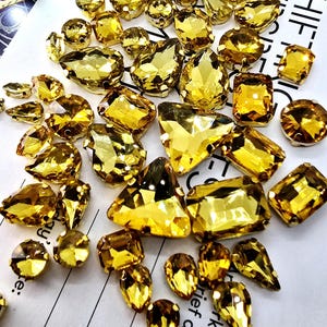 May include: A selection of golden-yellow rhinestones in diverse shapes, such as round, teardrop, and rectangular. The gemstones are scattered on a white surface, reflecting light. Suitable for jewellery making and craft projects. Various sizes and shapes.