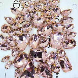 Può includere: Una collezione di strass di vetro rosa con montature dorate. Gli strass sono di varie forme e dimensioni.