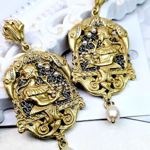 Può includere: Orecchini dorati ornati con un design a rilievo dettagliato con una figura classica, accentuati con smalto nero, pietre preziose blu e piccole perle. Ogni orecchino ha una perla pendente a forma di goccia.
