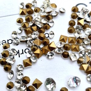 Può includere: Una spolverata di strass dorati e trasparenti. Gli strass sono di varie forme, tra cui rotonda, quadrata e piramidale. Gli strass sono sparsi su una superficie bianca.