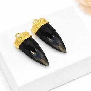 May include: Two black, faceted gemstone pendants with gold-colored caps and loops. The pendants are horn-shaped and sit on a white surface. A peach-colored rose is in the background.