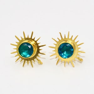 May include: A pair of gold-colored stud earrings. Each earring features a central, round, teal-colored gemstone surrounded by a sunburst design. The earrings are set against a white background.