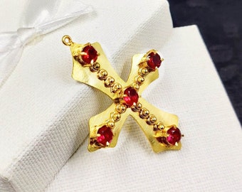 May include: A gold cross pendant with five red gemstones. The cross is detailed with a beaded border.