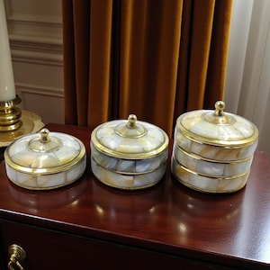 May include: Three decorative boxes with lids. The boxes are round with a mother-of-pearl mosaic design and gold-coloured trim. Each box has a small, round gold-coloured knob on top. The boxes are arranged on a dark wood surface.