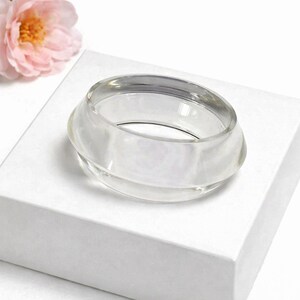 May include: A clear, circular glass bracelet sits on a white box, with a pink flower in the background. The bracelet has a rounded, layered design. The overall aesthetic is minimalist and elegant.