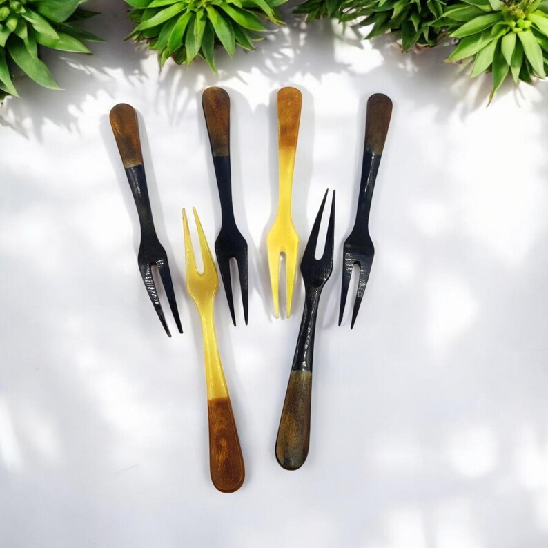 May include: A collection of six small forks with a two-pronged design. The forks have a two-tone color scheme, with black, yellow, and brown handles. The forks are arranged on a white surface, with green plants in the background.