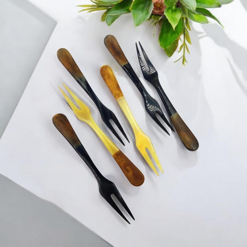 May include: A collection of six small forks with a two-pronged design. The forks have dark brown wooden handles and black or yellow tines. The forks are arranged on a white surface, with greenery in the background.