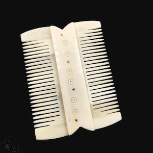 May include: Two ivory-coloured combs are joined at the centre. Each comb has a rounded top and a series of thin teeth. The connecting piece has decorative circles and small rivets. The combs are set against a black background.