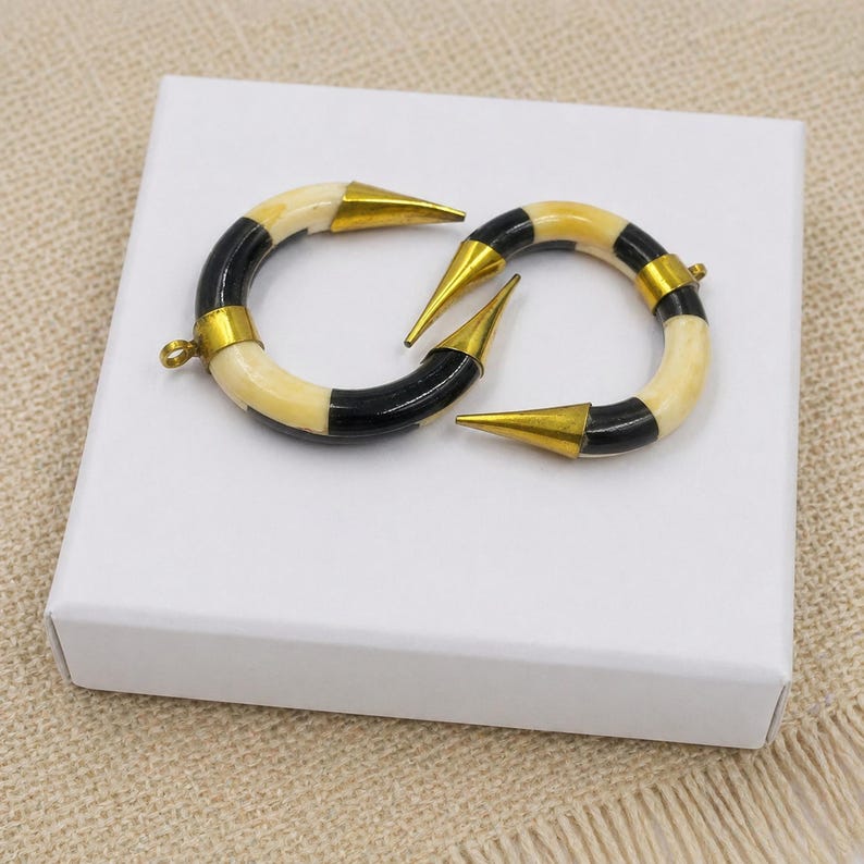 Peut inclure: Deux pendentifs en forme de croissant compos&eacute;s de segments altern&eacute;s de couleur cr&egrave;me et noire. Chaque pi&egrave;ce est agr&eacute;ment&eacute;e d'embouts en m&eacute;tal dor&eacute; poli aux deux extr&eacute;mit&eacute;s et comprend un petit anneau circulaire pour la fixation.