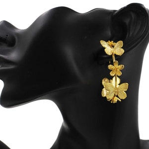 Puede incluir: Pendientes de aro dorados con una cadena y tres colgantes de mariposa. Las mariposas tienen detalles de alas texturizadas y un dise&ntilde;o floral.