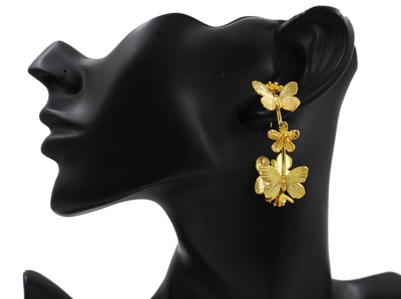 Puede incluir: Pendientes de aro dorados con una cadena y tres colgantes de mariposa. Las mariposas tienen detalles de alas texturizadas y un dise&ntilde;o floral.