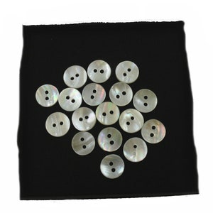 May include: A collection of 18 white mother-of-pearl buttons with two holes each. The buttons are arranged in a circular pattern on a black fabric background.