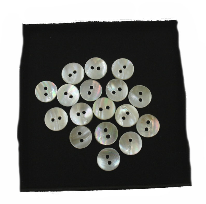 May include: A collection of 18 white mother-of-pearl buttons with two holes each. The buttons are arranged in a circular pattern on a black fabric background.