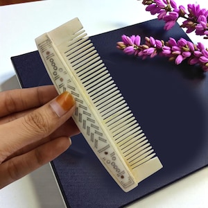 May include: Ivory-colored comb with intricate geometric and dot patterns. The comb's teeth are closely spaced, and the design includes small, dark accents. The comb is held against a dark blue surface, with a sprig of purple flowers in the background.