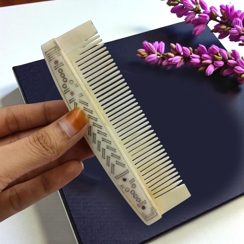 May include: Ivory-colored comb with intricate geometric and dot patterns. The comb's teeth are closely spaced, and the design includes small, dark accents. The comb is held against a dark blue surface, with a sprig of purple flowers in the background.