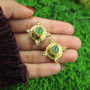 Puede incluir: Un par de pendientes de oro con detalles de piedras preciosas verdes. Los pendientes tienen un diseño circular con un borde decorativo.