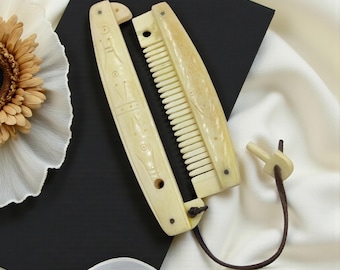 May include: An antique folding comb made of bone, with carved floral and geometric designs. The comb is off-white with a dark brown leather cord and a small wooden toggle. The comb is open, revealing the teeth.