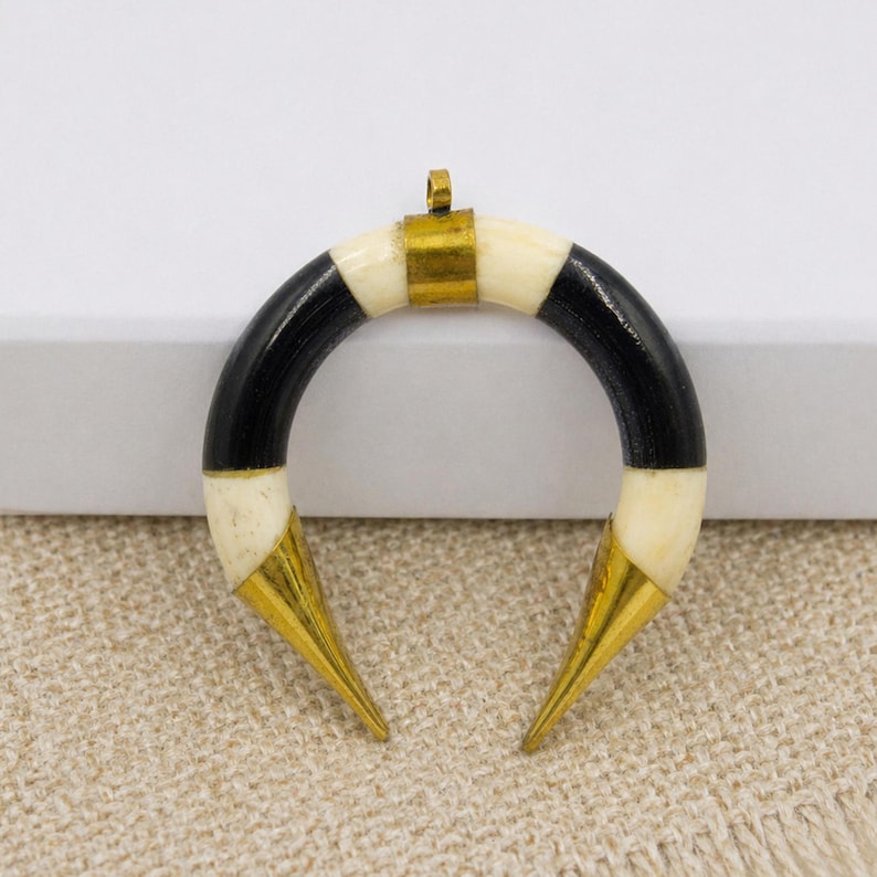 Peut inclure: Un pendentif en forme de croissant compos&eacute; de segments altern&eacute;s de mati&egrave;re lisse noire et blanc cass&eacute;. La pi&egrave;ce est agr&eacute;ment&eacute;e d'une b&eacute;li&egrave;re centrale en m&eacute;tal dor&eacute; au sommet et de pointes en m&eacute;tal dor&eacute; aux deux extr&eacute;mit&eacute;s de la courbe.