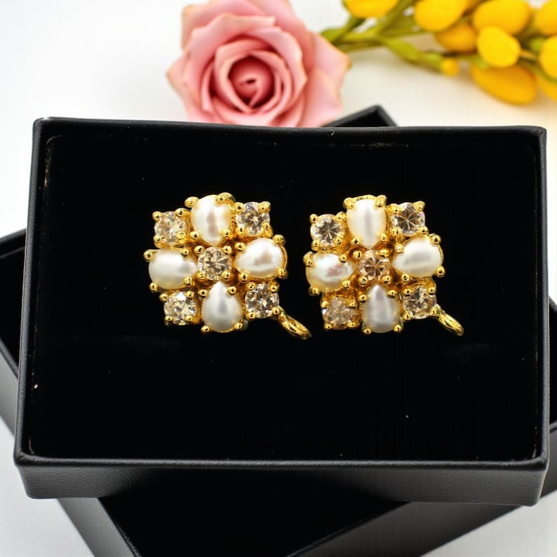 May include: A pair of gold-toned stud earrings with a floral design. Each earring features a cluster of white pearls and clear, sparkling crystals. The earrings are presented in a black jewelry box.