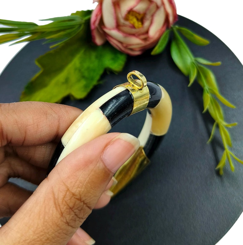 Peut inclure: Un bracelet jonc circulaire compos&eacute; de segments altern&eacute;s en mat&eacute;riau lisse noir et cr&egrave;me. Le bracelet est agr&eacute;ment&eacute; d'une pi&egrave;ce de connexion en m&eacute;tal dor&eacute; poli qui comprend une petite boucle int&eacute;gr&eacute;e sur la partie sup&eacute;rieure.