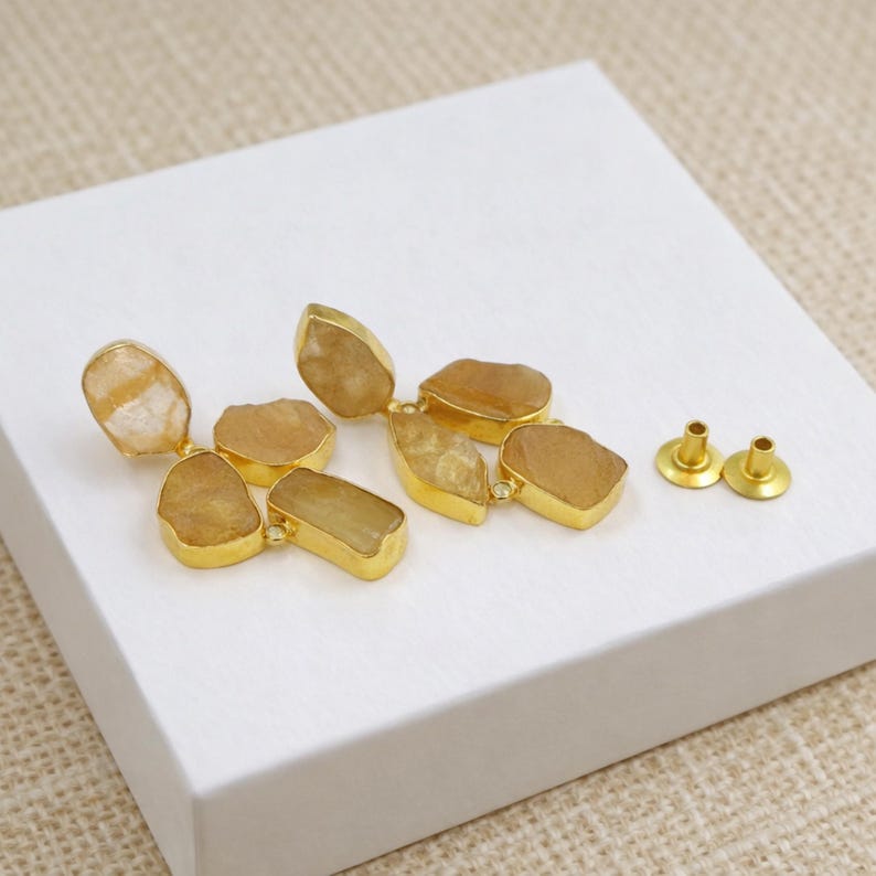 May include: A pair of gold-toned earrings with irregular, light brown stone accents. The earrings are composed of multiple stone shapes, each set in a gold frame. Two gold-toned earring posts are also visible.