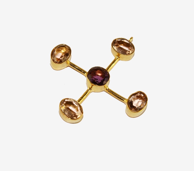 Puede incluir: Colgante dorado con dise&ntilde;o en forma de cruz. En el centro, una gema redonda de color p&uacute;rpura oscuro, rodeada por cuatro gemas ovaladas de color marr&oacute;n claro. El colgante se muestra sobre un fondo blanco.
