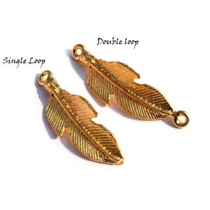 Pode incluir: Dois pingentes de pena dourados com estilos de laço diferentes. O pingente à esquerda tem um laço simples, enquanto o pingente à direita tem um laço duplo. Ambos os pingentes apresentam um design de pena detalhado.