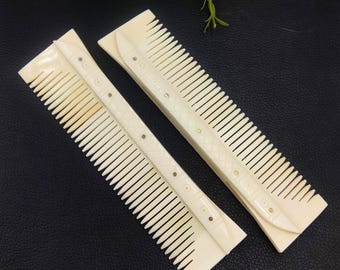 May include: Two off-white combs with fine teeth and decorative detailing. The combs have a rectangular shape with a slightly curved handle. Small gold-coloured studs adorn the handles. The combs are placed on a dark surface.