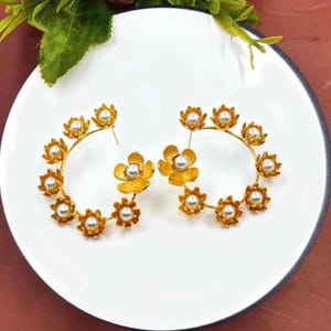 May include: A pair of gold hoop earrings with a floral design. Each earring has a circle of small gold flowers with white pearls in the center. The flowers are connected by a gold hoop. The earrings are on a white plate.