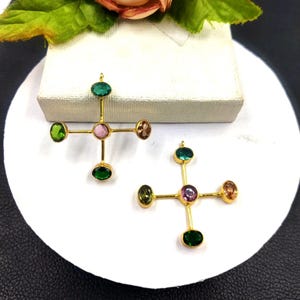 Puede incluir: Dos collares colgantes dorados con dise&ntilde;o de cruz. Cada cruz presenta cinco piedras preciosas ovaladas en tonos verdes, rosas y marrones. Los colgantes se exhiben sobre una superficie blanca.