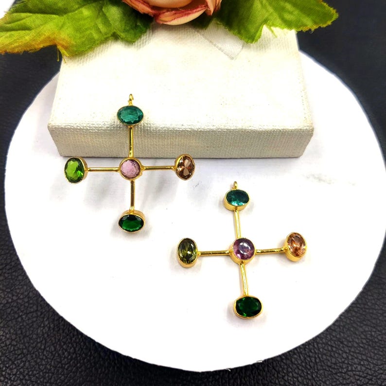 Puede incluir: Dos collares colgantes dorados con dise&ntilde;o de cruz. Cada cruz presenta cinco piedras preciosas ovaladas en tonos verdes, rosas y marrones. Los colgantes se exhiben sobre una superficie blanca.