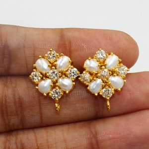 May include: A pair of gold-colored stud earrings. Each earring features a floral design with white teardrop-shaped pearls and clear, round-cut crystals. The earrings have a post and loop for attachment.
