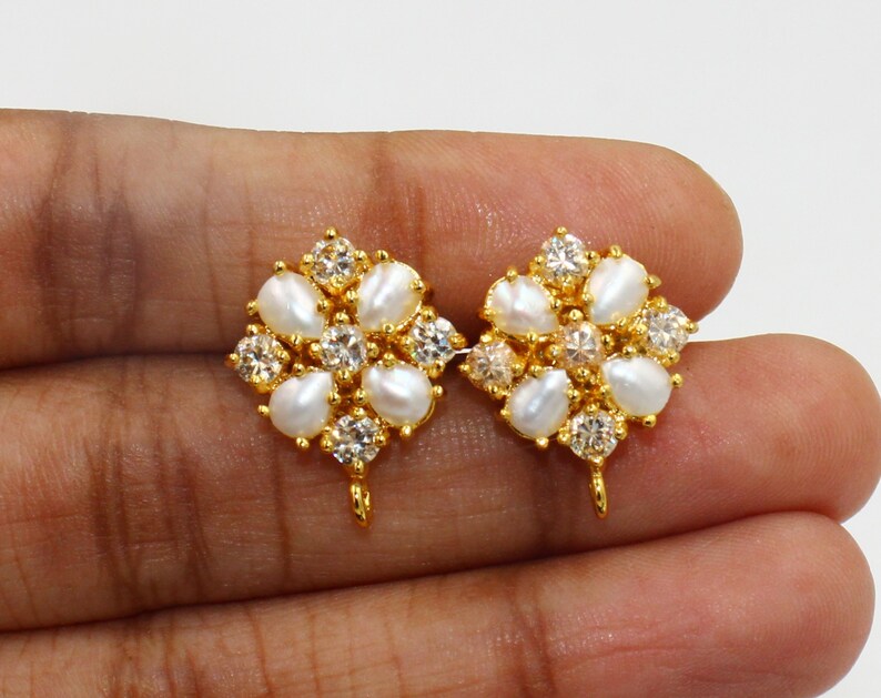May include: A pair of gold-colored stud earrings. Each earring features a floral design with white teardrop-shaped pearls and clear, round-cut crystals. The earrings have a post and loop for attachment.