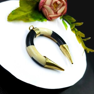 Peut inclure: Un pendentif incurv&eacute; compos&eacute; de segments altern&eacute;s de mati&egrave;re lisse couleur cr&egrave;me et noire. La pi&egrave;ce est dot&eacute;e d'embouts en m&eacute;tal dor&eacute; poli aux deux extr&eacute;mit&eacute;s pointues et d'une petite b&eacute;li&egrave;re circulaire au sommet pour la fixation.