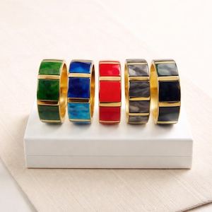 May include: Five colourful bangles with gold accents are displayed on a white surface. The bangles feature green, blue, red, grey, and black sections. Each bangle has a gold band in the middle and at the top and bottom.