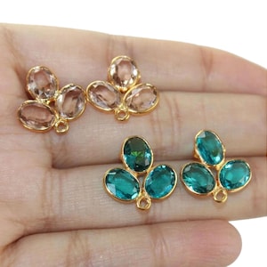 May include: Two pairs of gold-tone earring charms with three oval-shaped crystal stones. One pair features light pink stones and the other pair features teal blue stones.