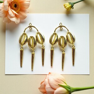 Könnte beinhalten: Ein Paar goldfarbene Ohrringe mit Muscheldesign und drei konischen Anhängern. Die Ohrringe werden auf einer weißen Oberfläche präsentiert, mit pfirsichfarbenen Blumen im Hintergrund.