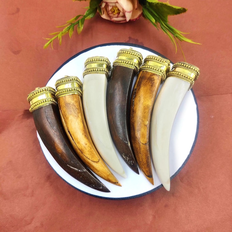 May include: Seven wooden pendants in the shape of a canine tooth, with gold-colored metal accents. The pendants are arranged in a semi-circle on a white plate.