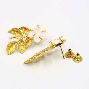 May include: A pair of gold-toned floral earrings with a white flower and leaf design. The earrings have a post and backing for secure wear. The earrings are set against a white background.