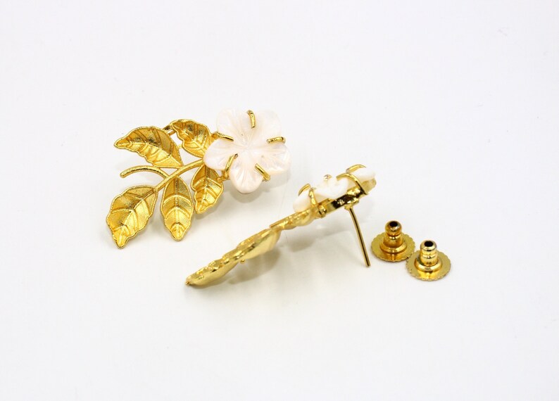 May include: A pair of gold-toned floral earrings with a white flower and leaf design. The earrings have a post and backing for secure wear. The earrings are set against a white background.