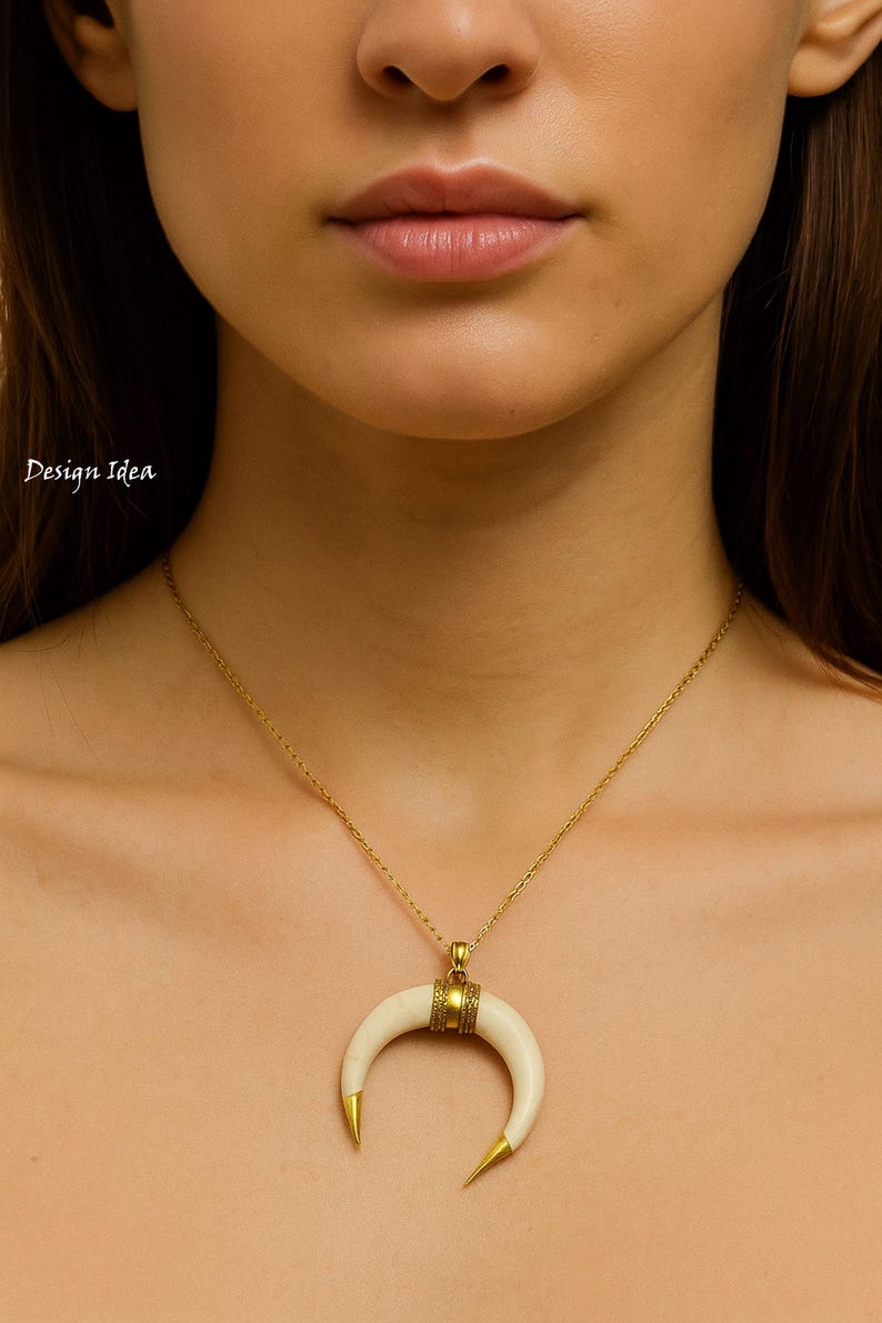 May include: A gold necklace featuring a crescent moon pendant. The pendant is off-white with gold tips and a gold band. The necklace is worn around the neck.