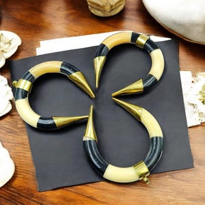 Peut inclure: Trois bracelets incurv&eacute;s en forme de croissant avec des segments altern&eacute;s de couleur cr&egrave;me et noire. Chaque bracelet est orn&eacute; d'embouts en m&eacute;tal dor&eacute; poli aux extr&eacute;mit&eacute;s pointues et d'une petite boucle m&eacute;tallique pour la fixation.
