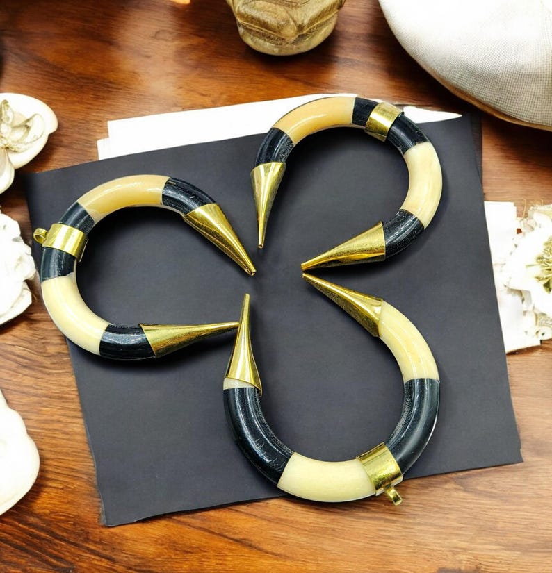 Peut inclure: Trois bracelets incurv&eacute;s en forme de croissant avec des segments altern&eacute;s de couleur cr&egrave;me et noire. Chaque bracelet est orn&eacute; d'embouts en m&eacute;tal dor&eacute; poli aux extr&eacute;mit&eacute;s pointues et d'une petite boucle m&eacute;tallique pour la fixation.
