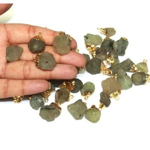 Könnte beinhalten: Eine Sammlung von roh geschnittenen, olivgrünen Edelsteinanhängern mit goldfarbenen Metallkappen und Schlaufen. Die Anhänger variieren in Größe und Form und haben ein natürliches, organisches Aussehen.