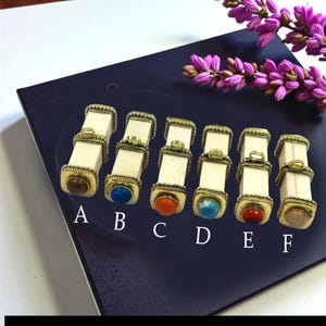 Può includere: Sei oggetti decorativi rettangolari color avorio con dettagli in metallo dorato e pietre preziose colorate. Le pietre sono marroni, blu, arancioni, turchesi, rosse e rosa. Etichettati dalla A alla F.