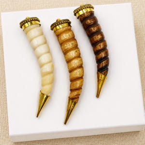 May include: Three curved, horn-shaped decorative objects with a spiral texture. Each piece features a metallic gold-toned cap at the top with a beaded edge and a smooth, pointed gold-toned tip at the bottom. The bodies are cream, tan, and dark brown.