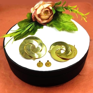 May include: A pair of gold-tone earrings with a twisted, circular design. The earrings are displayed on a white box with a black lid. Two earring backs are also included.
